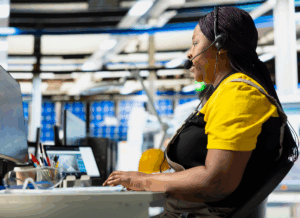 A woman wearing a headset works at a computer station inside a modern manufacturing facility, symbolizing the integration of communication and technology in the manufacturing sales process.