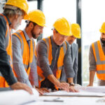 A construction project manager at a desk reviewing blueprints side-by-side with financial reports on a laptop/tablet.