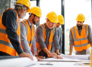 A construction project manager at a desk reviewing blueprints side-by-side with financial reports on a laptop/tablet.