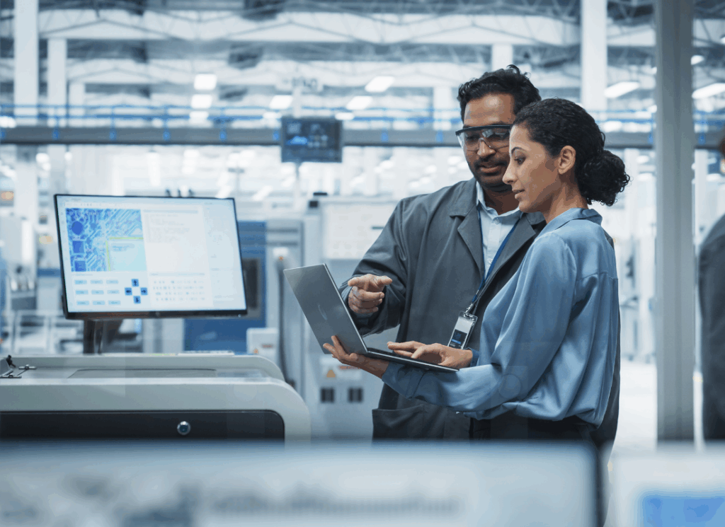 Two engineers review data on a laptop in a factory, symbolizing the importance of cybersecurity in manufacturing operations.