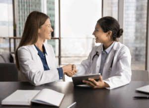 Two female doctors smiling and shaking hands in an office, and collaborating on starting a medical practice.