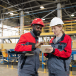 Two factory workers in hard hats review data on a tablet, symbolizing cost reduction strategies in manufacturing.