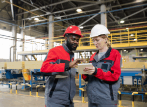 Two factory workers in hard hats review data on a tablet, symbolizing cost reduction strategies in manufacturing.