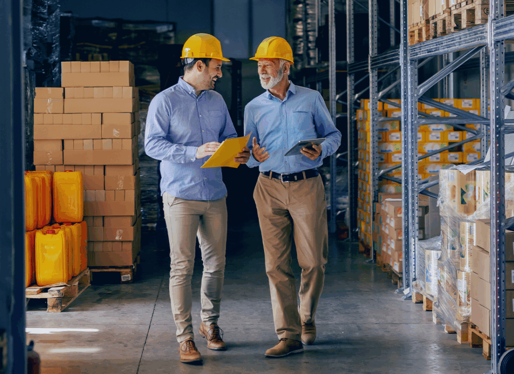 Two business partners in formal wear and with protective yellow helmets on heads walking and talking about business. Younger one holding folder with data while older one using tablet.