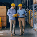 Two business partners in formal wear and with protective yellow helmets on heads walking and talking about business. Younger one holding folder with data while older one using tablet.