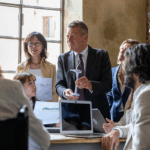 A group of six business professionals gathers around a table in a meeting room with natural light coming through a large window. One man in a suit is holding a small model of a wind turbine while others listen attentively. Laptops and printed charts are visible on the table, suggesting a discussion about renewable energy or sustainability.