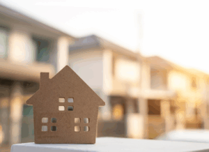 A small wooden house model sits in front of blurred homes in warm sunlight, representing energy-efficient buildings qualifying for the 179D deduction.