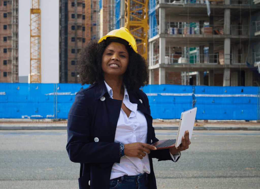 A construction accountant wearing a hard hat reviews information on a laptop at a job site, representing construction income statements..