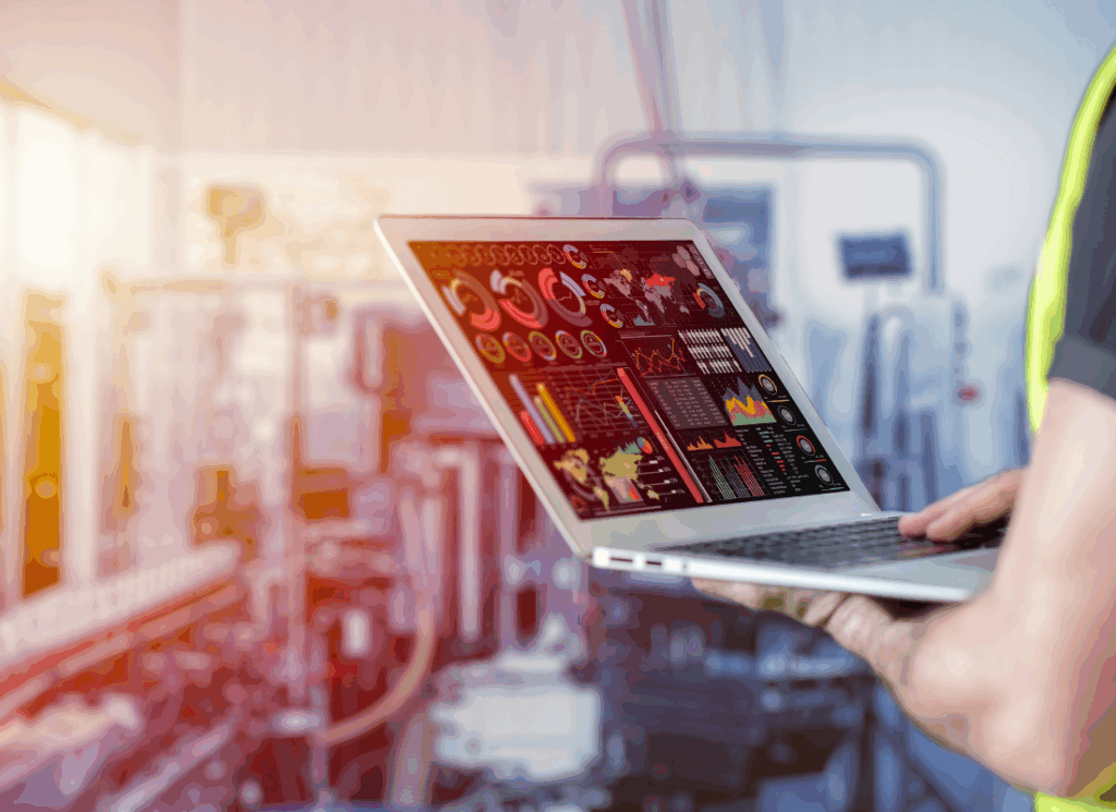 A person in a safety vest reviews real-time production dashboards on a laptop inside a manufacturing facility, illustrating insights for a manufacturing pricing strategy.