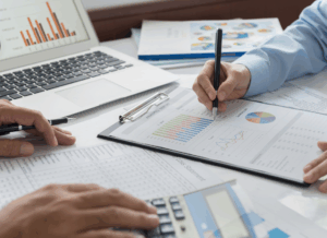 Two dental accounting professionals review financial charts and reports with a laptop and calculator on the table, illustrating detailed analysis for Dental Business Consulting.