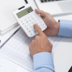 A CPA using a calculator over financial spreadsheets at a desk, reviewing figures related to a manufacturing chart of accounts.