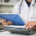 A doctor reviews paperwork while using a calculator and laptop, highlighting financial planning and Tax benefits for doctors in a clinical office setting.