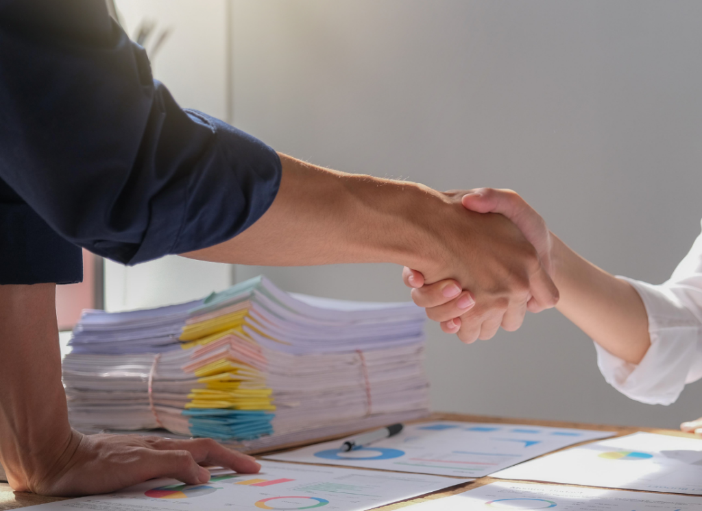 Close-up of two people shaking hands over financial documents with charts and a large stack of paperwork on a desk.