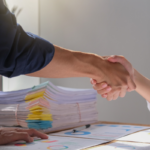 Close-up of two people shaking hands over financial documents with charts and a large stack of paperwork on a desk.