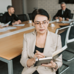 A business professional taking notes in a meeting while colleagues discuss documents at a conference table in the background.