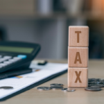 Wooden blocks spelling “TAX” stacked on a desk with coins, a calculator, and financial documents in the background.