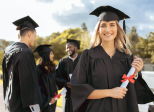 Smiling graduate in cap and gown holding a diploma with classmates in the background during an outdoor graduation ceremony.