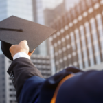 Graduate in cap and gown holding up a mortarboard against a city skyline at sunset.