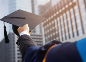 Graduate in cap and gown holding up a mortarboard against a city skyline at sunset.