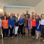 Group photo of the James Moore team standing together indoors, smiling and dressed in business casual attire during a team gathering.