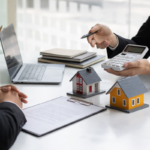 Two professionals reviewing documents as one uses a calculator beside model homes and a laptop on a desk.