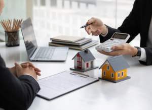 Two professionals reviewing documents as one uses a calculator beside model homes and a laptop on a desk.