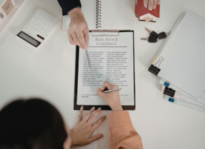 Overhead view of a person signing a real estate contract on a clipboard, with house keys, a calculator, and office supplies on the desk.