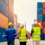 Three logistics professionals wearing hard hats and high-visibility vests walk through a shipping container yard, discussing operations and pointing toward stacked cargo containers.