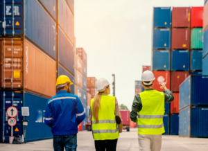 Three logistics professionals wearing hard hats and high-visibility vests walk through a shipping container yard, discussing operations and pointing toward stacked cargo containers.