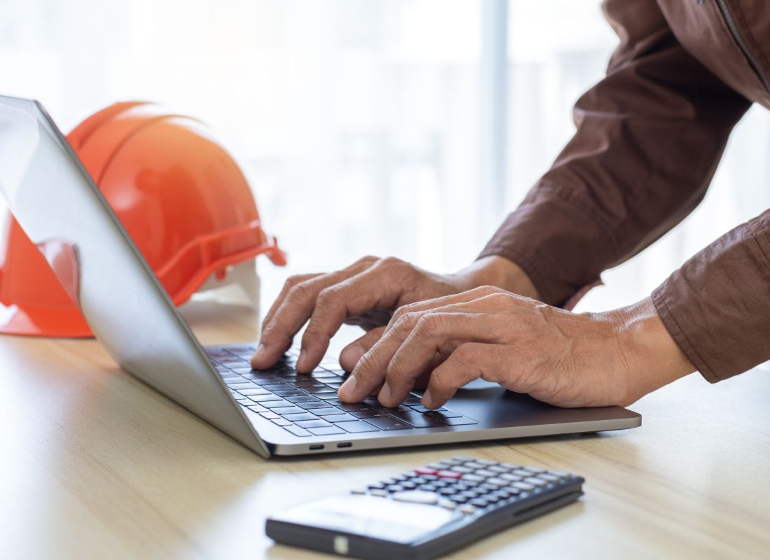 Construction professional using a laptop for project budgeting and financial planning with a hard hat and calculator on a desk.