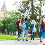 Group of students with backpacks walking together on a college campus pathway surrounded by trees and historic buildings.