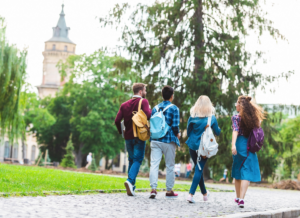 Group of students with backpacks walking together on a college campus pathway surrounded by trees and historic buildings.