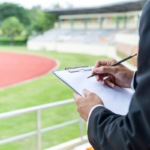 Business professional holding a clipboard and taking notes while overlooking an outdoor athletic track and stadium seating.