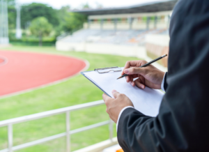 Business professional holding a clipboard and taking notes while overlooking an outdoor athletic track and stadium seating.