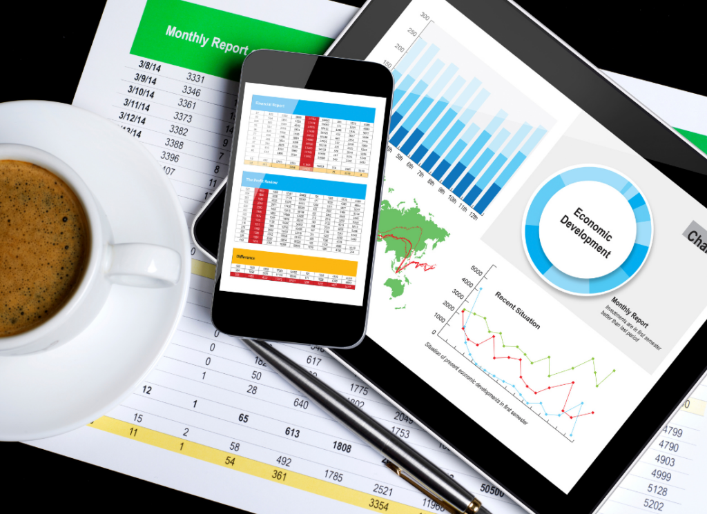 Tablet and smartphone displaying business reports and financial charts on a desk with printed spreadsheets, a pen, and a cup of coffee.