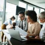 Group of business professionals collaborating in an office while reviewing documents and financial reports around a laptop.