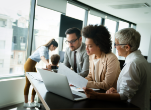 Group of business professionals collaborating in an office while reviewing documents and financial reports around a laptop.