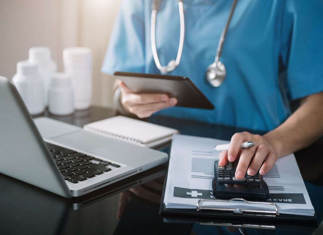 A healthcare professional in blue scrubs uses a tablet while calculating costs on a calculator beside a laptop and medical paperwork in a clinical office.