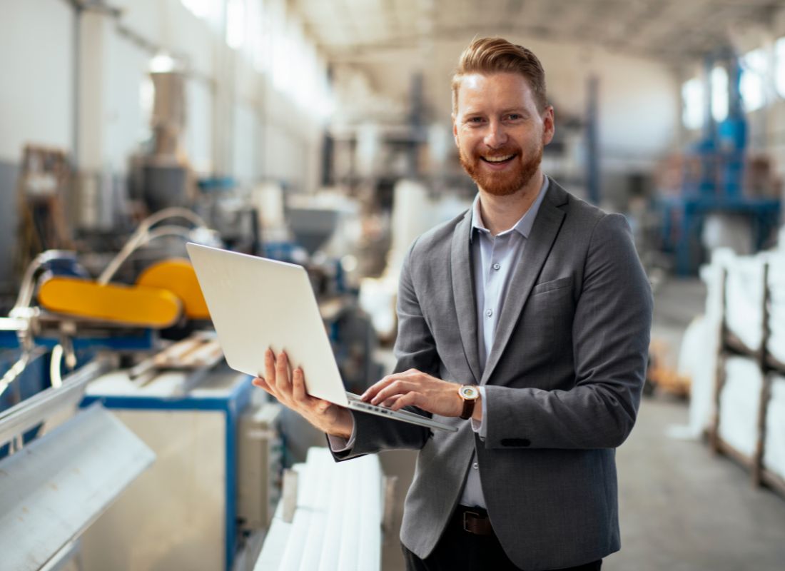 A manufacturing professional in business attire uses a laptop while standing on a factory floor, with industrial equipment and production machinery in the background.