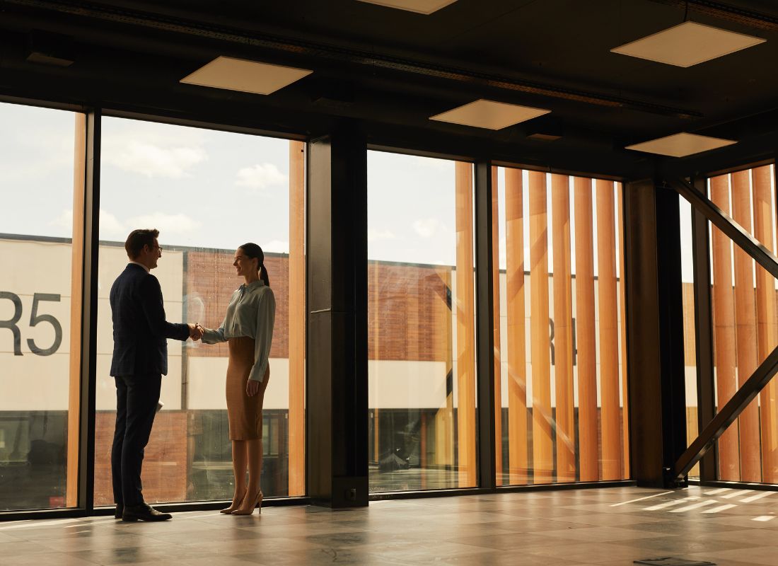Two commercial real estate professionals shake hands in a modern office with floor-to-ceiling windows, symbolizing a successful property transaction, lease agreement, or business partnership.