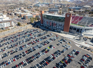 Aerial view of a college football stadium beside a large parking lot filled with cars, with university buildings and mountains visible in the background.