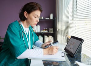 Physician in scrubs taking notes while checking her phone, with a video call open on a tablet at her desk.