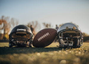 Two football helmets and a football resting on a grass field at sunset, with blurred figures in the background.