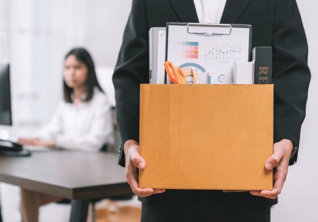 Are Layoffs Looming featured image 1100 x 800 (80) An employee is in an office holding a box of office supplies and personal items. In the background, a person is at a desk on a computer.