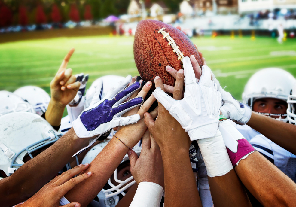 A football team huddles on the field, with players in helmets and gloves reaching toward the center and raising a football above their hands.