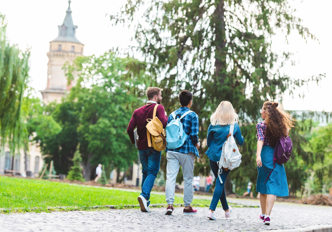 Group of students with backpacks walking together on a college campus pathway surrounded by trees and historic buildings.