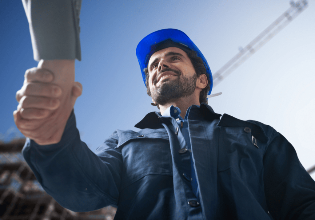 A construction worker is smiling and shaking hands with a person who is mostly off camera, aside from their arm. The construction worker appears to be a new hire shaking hands with his employer. The background shows part of a building and construction equipment.