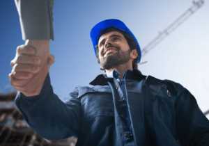 Benefits of Robust Onboarding featured image 1100 x 800 A construction worker is smiling and shaking hands with a person who is mostly off camera, aside from their arm. The construction worker appears to be a new hire shaking hands with his employer. The background shows part of a building and construction equipment.