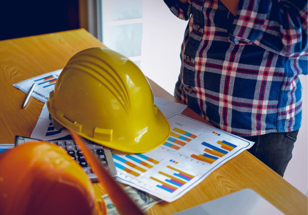 Connecting Operations With Finance featured image Two hard hats are on a desk in an office next to a calculator and financial reports. A person is standing over the desk.