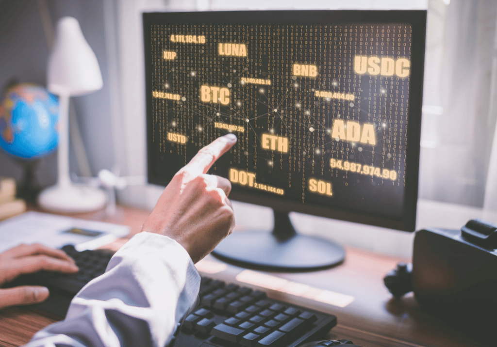 Cryptocurrency Basics featured image Someone is pointing to a computer monitor on a desk. The monitor is displaying various cryptocurrency names.